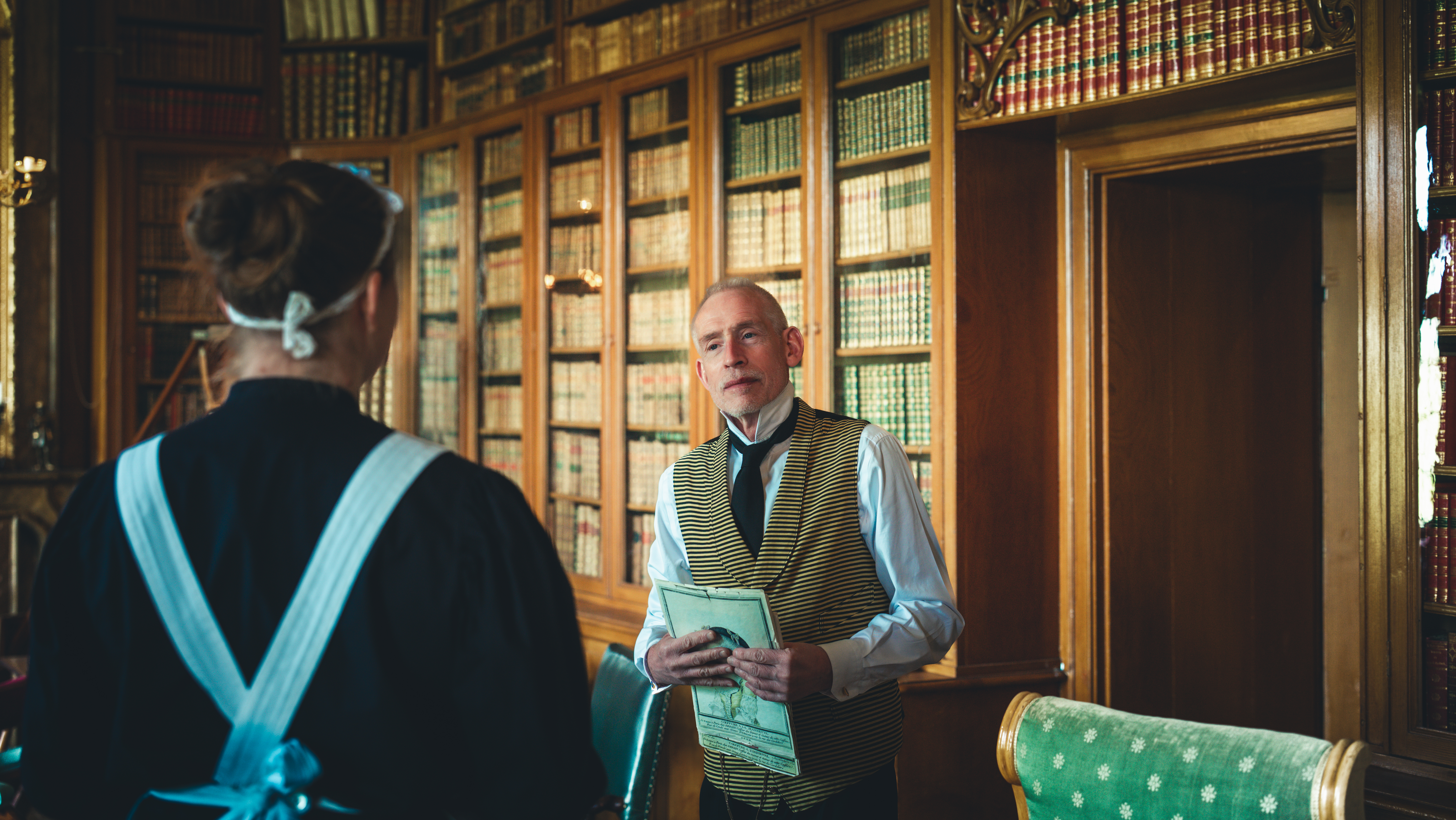 Betjänten Wilhelm i dialog med husan Ida i biblioteket på Löfstad slott.