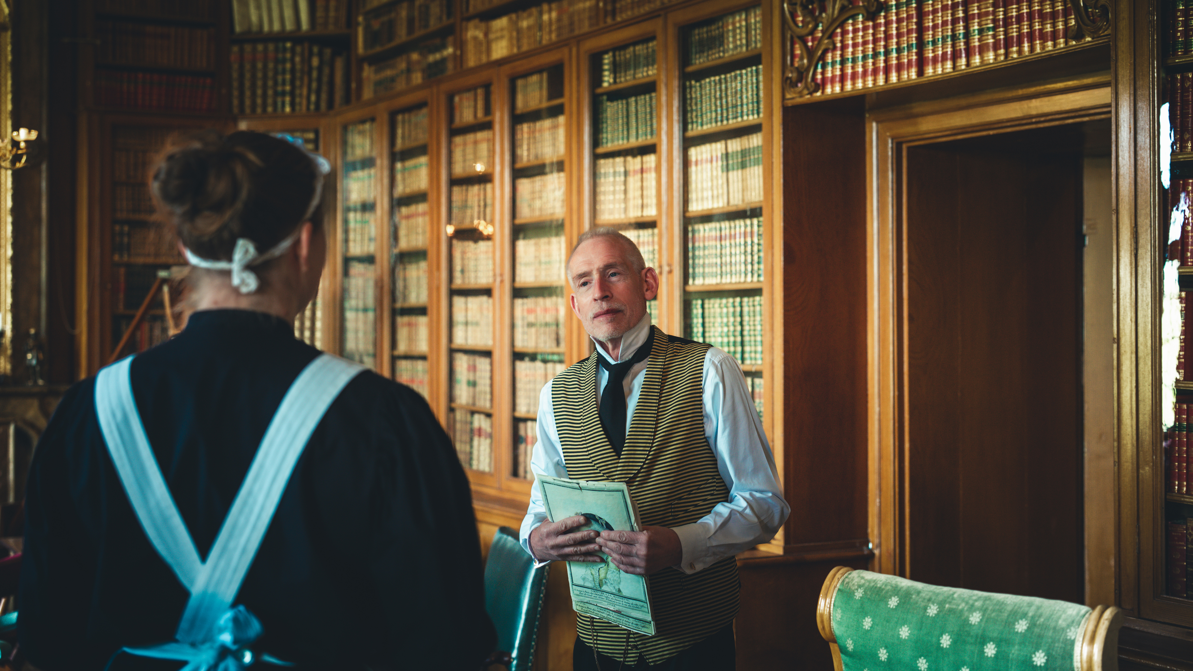 Betjänten Wilhelm i dialog med husan Ida i biblioteket på Löfstad slott.